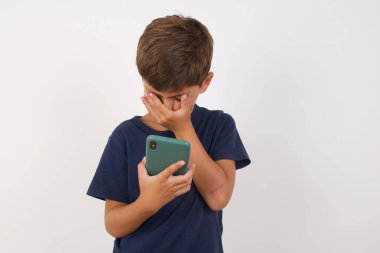 Güzel çocuk günlük tişört giyip izole edilmiş beyaz arka planda dikiliyor. Akıllı telefona bakıyor ve üzgün bir şekilde yüz yüze bakıyor..