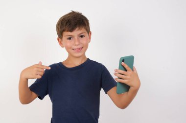 Neşeli, çekici, sıradan tişörtler giyen güzel çocuk izole beyaz arka planda duruyor, elinde telefonla yeni bir elma telefonu modeli var.