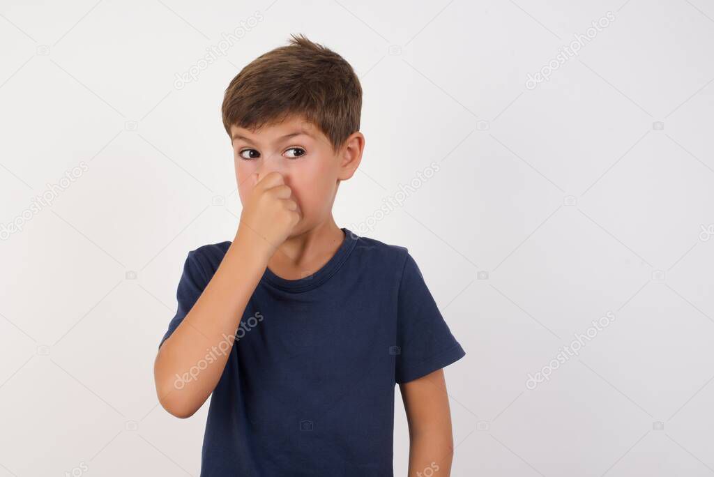 Beautiful kid boy wearing casual t-shirt standing over isolated white background,smelling something stinky and disgusting, intolerable smell, holding breath with fingers on nose. Bad smell