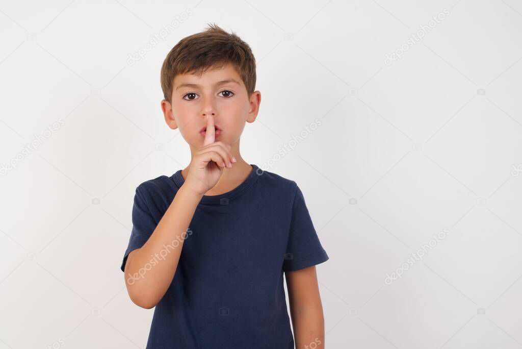 Beautiful kid boy wearing casual t-shirt standing over isolated white background, asking to be quiet with finger on lips. Silence and secret concept.