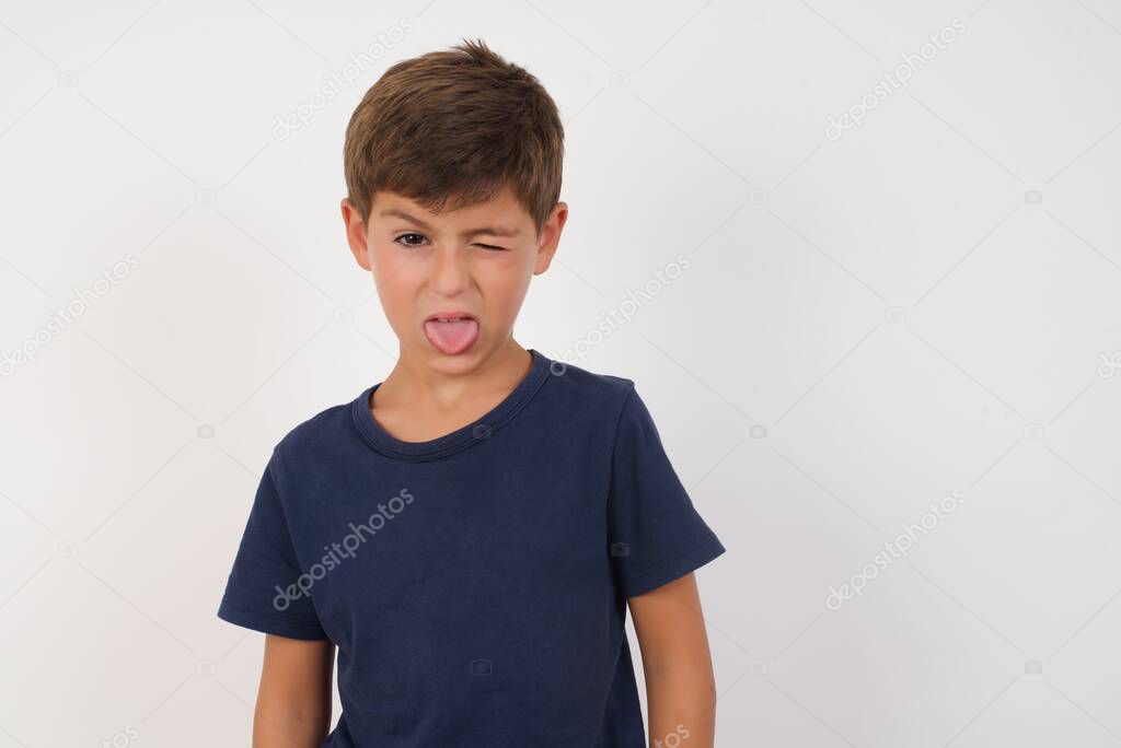 Beautiful kid boy wearing casual t-shirt standing over isolated white background, sticking tongue out happy with funny expression. Emotion concept.