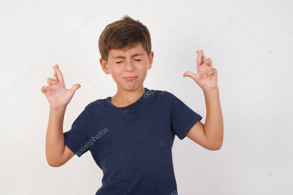Beautiful kid boy wearing casual t-shirt standing over isolated white background, gesturing finger crossed smiling with hope and eyes closed. Luck and superstitious concept.