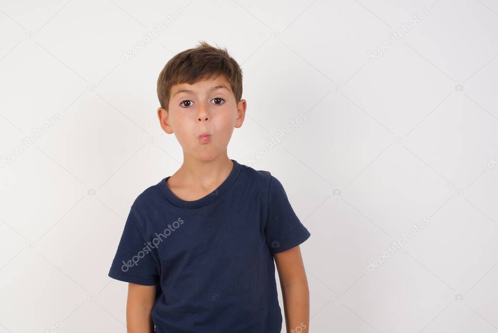 Shot of pleasant looking brunette Beautiful kid boy wearing casual t-shirt standing over isolated white background, pouts lips, looks  at camera,   Human facial expressions