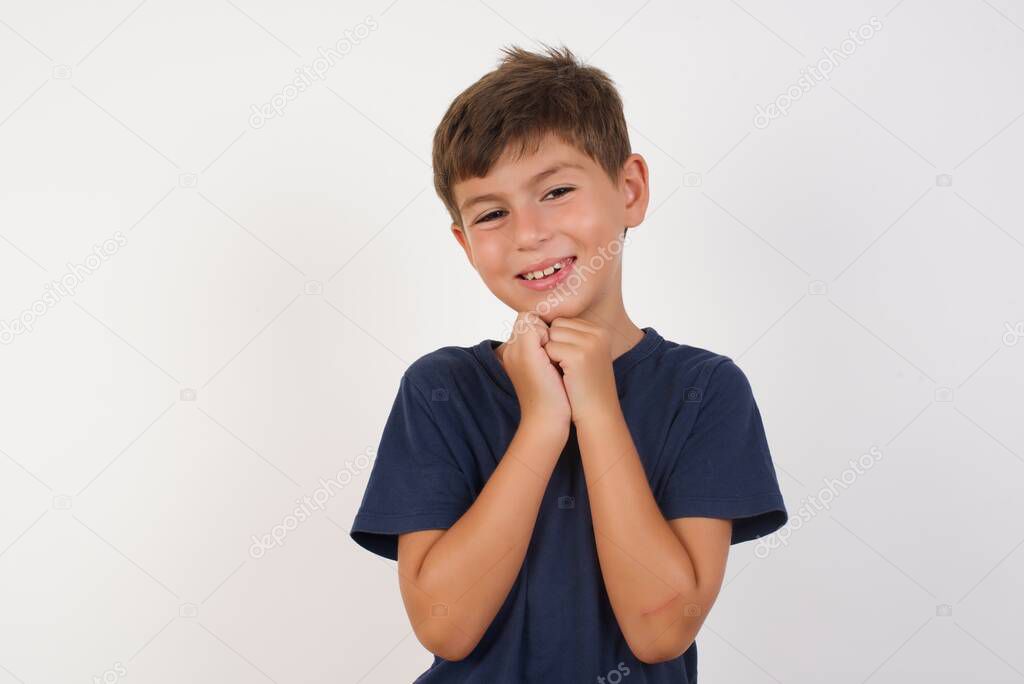 Positive adorable Beautiful kid boy wearing casual t-shirt standing over isolated white background, smiles happily, glad to receive pleasant news from interlocutor, keeps hands together. People emotions concept.