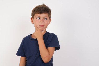 Güzel bir çocuk, izole edilmiş beyaz arka planda duran sıradan bir tişört giyer ve bir soru hakkında endişelenir, eli çenesinde endişeli ve gergindir..