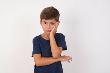 Çok sıkılmış, rahat tişört giyen güzel çocuk izole beyaz arka planda dikiliyor. Bir yandan el ele tutuşurken, diğer yandan diğer yandan kollar çapraz, yorgun ve hasta görünüyor.,  