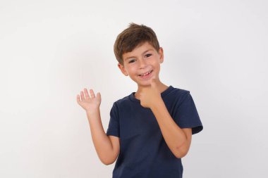 Güzel çocuk, izole edilmiş beyaz arka planda duran sıradan bir tişört giyiyor, avuç içini gösteriyor ve baş parmaklarıyla iyi bir hareket yapıyor, mutlu ve neşeli gülümsüyor..