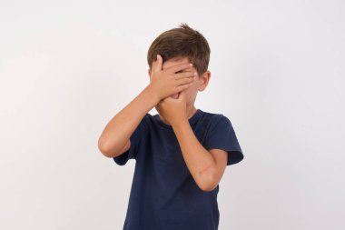 Güzel çocuk, izole edilmiş beyaz arka planda dikilen, ağzı ve gözlerini kapayan, şaşırmış ve şok olmuş bir tişört giyiyor. Duyguları gizlemek.