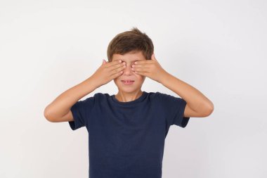 Güzel bir çocuk, izole edilmiş beyaz arka plan üzerinde sıradan bir tişört giyer, elleri gülücüklü ve komik bir şekilde gözlerini kapatır. Kör kavram.