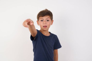 Güzel bir çocuk, izole edilmiş beyaz arka plan üzerinde duran sıradan bir tişört giyer, parmağıyla ileriyi işaret eder, ağzı açık ve şaşırmış bir ifade takınır..
