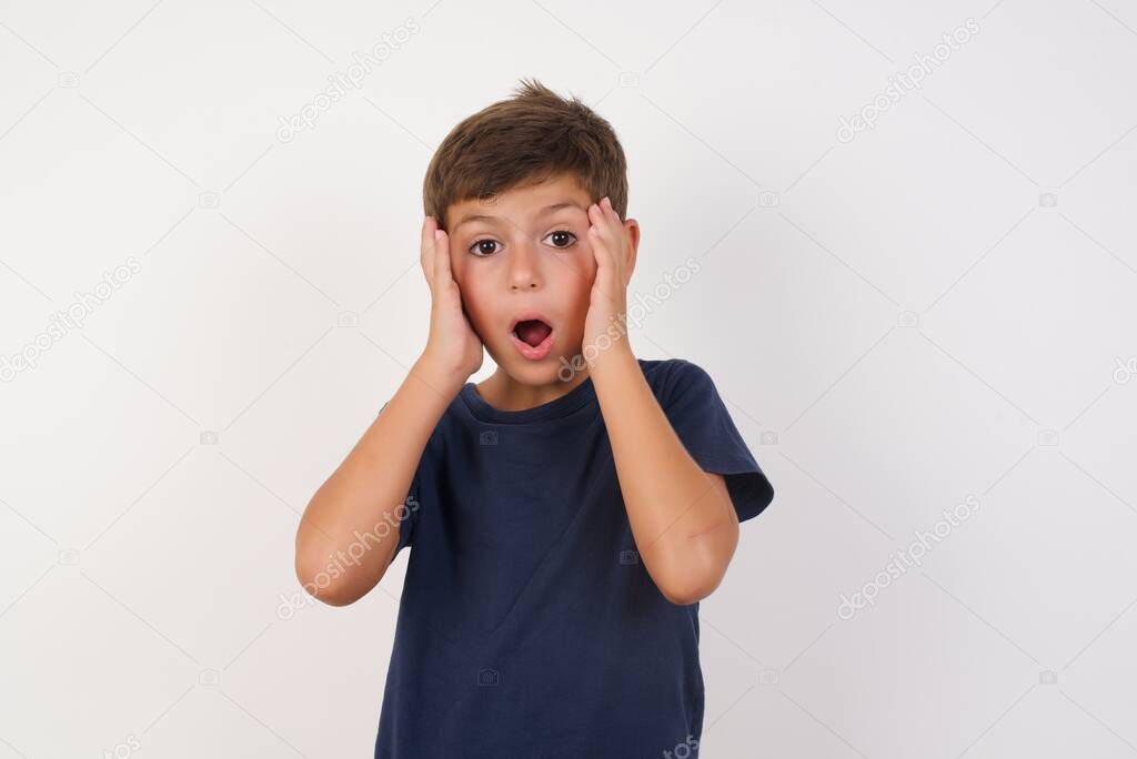 Emotive Beautiful kid boy wearing casual t-shirt standing over isolated white background, with scared expression, keeps hands on head, jaw dropped, has terrific expression. Omg concept