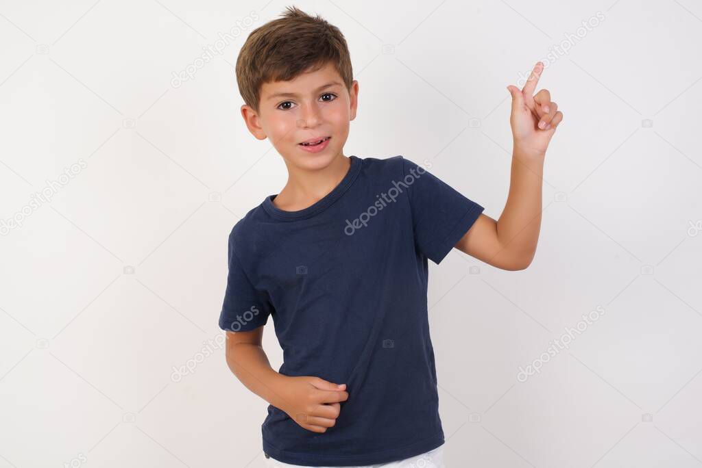 Smiling Beautiful kid boy wearing casual t-shirt standing over isolated white background, pointing up and looking at the camera 