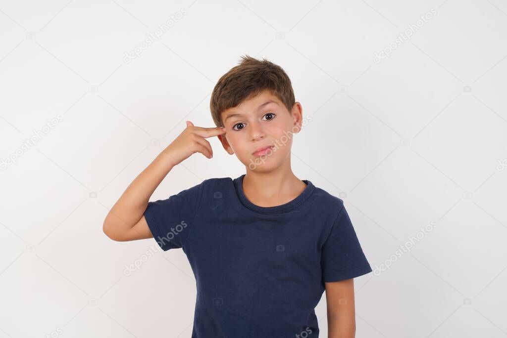Unhappy Beautiful kid boy wearing casual t-shirt standing over isolated white background,  makes suicide gesture and imitates gun with hand, curves lips, keeps two fingers on temple