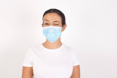 Tıbbi maske takan genç bir Arap kadın izole edilmiş beyaz arka planda durup mutlu bir şekilde gülümsüyor kameraya şakacı bir şekilde göz kırpıyor ve seninle flört ediyor. İnsan yüz ifadeleri ve duyguları