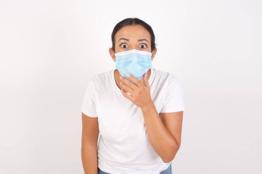 Kafası karışmış, tıbbi maske takan genç bir kadın izole edilmiş beyaz arka planda dikiliyor, şaşkınlıktan ağzı açılıyor, ani haberlere tepki veriyor. İnsanlar ve yüz ifadeleri.