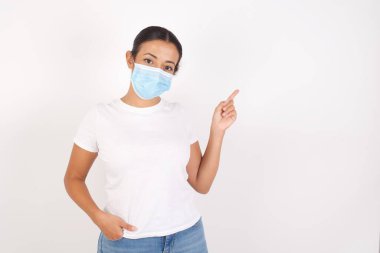 İzole edilmiş beyaz arka planda duran tıbbi maske takan genç bir Arap kadını işaret eden parmaklar boş yere bakıyor.