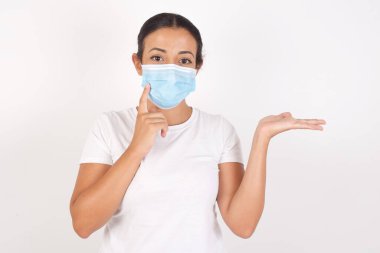 İzole edilmiş beyaz arka planda duran tıbbi maske takan komik genç Arap kadın yeni bir ürünü açık havada tutuyor. Harika bir öneri. 