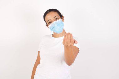 Tıbbi maske takan genç bir Arap kadın izole edilmiş beyaz arka planda duruyor ve elinden geleni yapmaya davet ediyor. Gelmene sevindim.