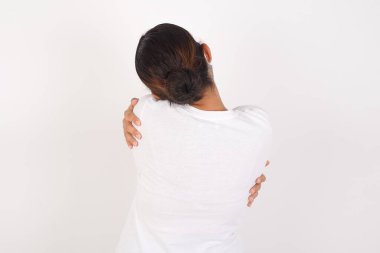Tıbbi maske takan genç bir Arap kadın izole edilmiş beyaz arka planda dikiliyor mutlu ve pozitif bir şekilde kendini kucaklıyor. Sevgi ve özgüven.