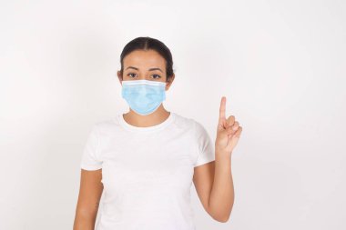 İzole edilmiş beyaz arka planda duran tıbbi maske takan genç Arap kadın bir numaralı parmağı gösterip işaret ederken kendinden emin ve mutlu bir şekilde gülümsüyor..