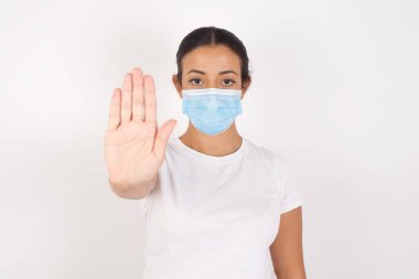 Tıbbi maske takan genç bir Arap kadın izole edilmiş beyaz arka planda duruyor ve avuç içiyle el hareketi yapıyor. Yüzünde olumsuz ve ciddi bir jest olan uyarı ifadesi 