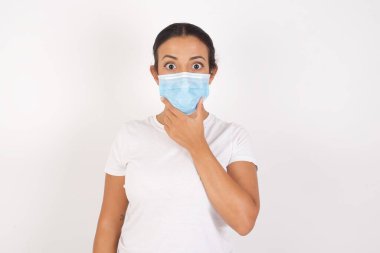 İzole edilmiş beyaz arka planda duran tıbbi maske takan genç bir Arap kadın elleri çenesinde olan inançsızlığa, şaşkınlığa ve hayrete düşmüş bir ifadeye bakıyor.