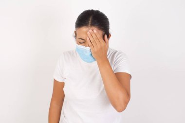 Tıbbi maske takan genç bir Arap kadın izole edilmiş beyaz arka planda duruyor. Esnemekten yorulmuş, yarı yüzü, gözü ve ağzı elle kapatıyor. Yüzüm acıyor..