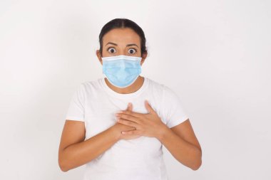 Korkak genç Arap kadın, tıbbi maske takıyor, izole edilmiş beyaz arka planda duruyor, korkmuş bir ifadeyle, ellerini göğsünde tutuyor, garip bir şey fark etmek için şaşırıyor, insanlar, sessiz tepki ve duygular..