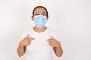 Genç Arap kadının tıbbi maske takıp vücudunun izole edilmiş beyaz arka plan noktalarının başında dikilirken, alışverişe çıktıktan sonra iyi bir ruh hali içinde olduğu ve başarılı alışverişler yaptığı yatay bir görüntü.