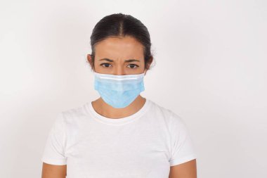İzole edilmiş beyaz arka planda duran tıbbi maske takan öfkeli genç Arap kadın portresi dişlerini sıkar, tatminsizlikten kaşlarını çatar, birçok görev yüzünden tedirgin olur. Öfkeli adam pencereden dışarı bakar, sefil havayı görür.