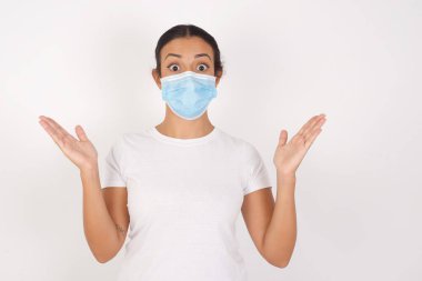 Ürkmüş genç Arap kadın tıbbi maske takıyor. İzole edilmiş beyaz arka plan hareketlerinin başında belirsizlikle duruyor. Kameraya bakıyor.