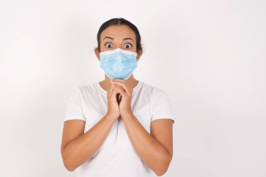 Tıbbi maske takan genç bir Arap kadın izole edilmiş beyaz arka planda durup şans dilemek için dua ediyor. Elleri çapraz, hayrete düşmüş ve ağzı açık bir şekilde öne bakıyor..