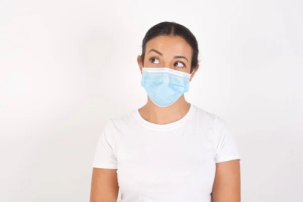 Amazed Young arab woman wearing medical mask standing over isolated white background bitting lip and looking up to empty space,