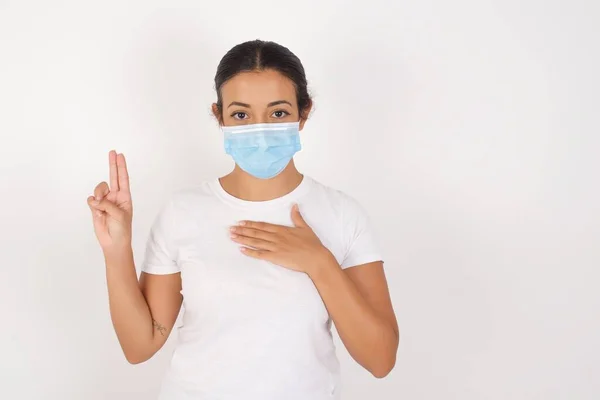 年轻的阿拉伯女人戴着医疗面具 站在与世隔绝的白色背景上 微笑着 双手托着胸膛和手指发誓 宣誓效忠 — 图库照片