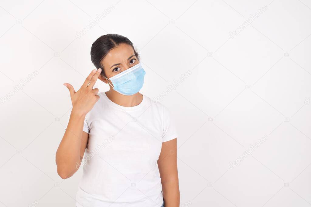 Unhappy  Young arab woman wearing medical mask standing over isolated white background  makes suicide gesture and imitates gun with hand, curves lips, keeps two fingers on temple