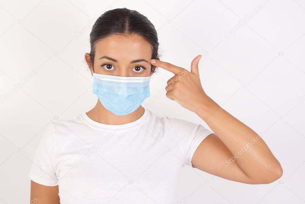 Unhappy Young arab woman wearing medical mask standing over isolated white background makes suicide gesture and imitates gun with hand, curves lips keeps two fingers on temple, shoots, being tired of everything,
