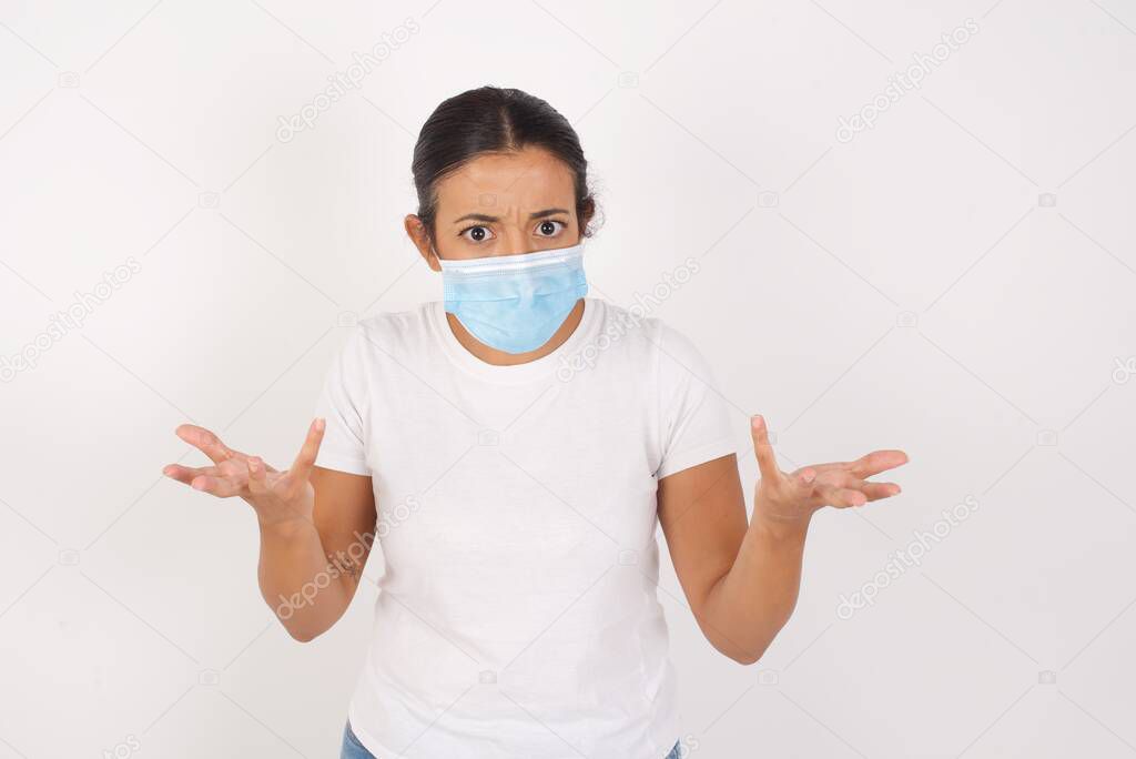 Frustrated Young arab woman wearing medical mask standing over isolated white background feels puzzled and hesitant, shrugs shoulders in bewilderment, keeps mouth widely opened, doesn't know what to do.