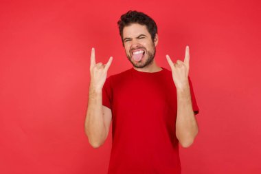 Stüdyoda poz veren esmer bir genç. Taş el hareketi yapıyor ve dil gösteriyor.