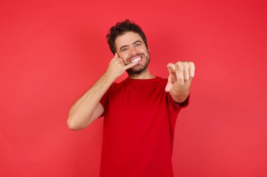 Genç, güzel, esmer, kırmızı tişörtlü, neşeli bir şekilde gülümseyen ve kamerayı işaret eden bir adam. 