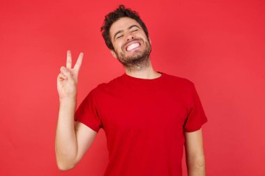 Kırmızı arka plan, stüdyoda gülen bir adam kameraya göz kırparak zafer işareti yapıyor. İki numara.