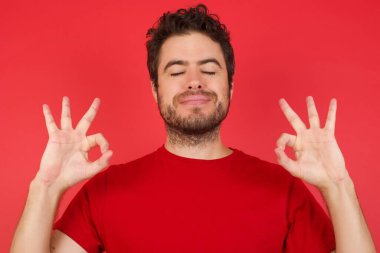 Beyaz arka plan üzerine gündelik kırmızı gömlek giyen genç, güzel esmer adam rahatla ve parmaklarıyla meditasyon hareketi yaparken gözleri kapalı gülümse. Yoga konsepti.