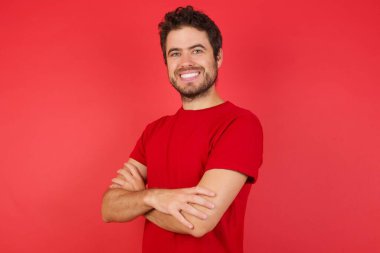 Kıvırcık saçlı, kırmızı tişörtlü, izole edilmiş sarı arka planda mutlu yüzlü, kollarını kavuşturup kameraya bakan genç bir Arap. Pozitif kişi.