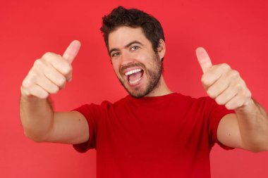 Kırmızı stüdyodaki beyaz adam, gündelik kırmızı gömlek giyiyor. Elle olumlu jest yapmayı onaylıyor. Başparmağıyla gülümsüyor ve başarı için seviniyor.