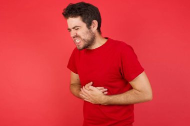 İzole edilmiş arka planda kırmızı tişört giyen yakışıklı genç bir adam mide bulantısı, acı verici bir hastalık yüzünden el üstünde tutuluyor. Ache kavramı.