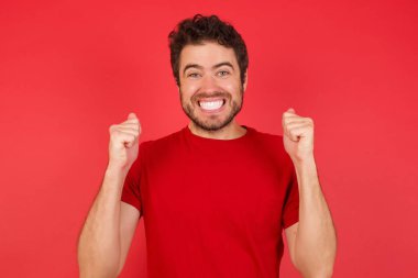 İzole edilmiş kırmızı arka plan üzerinde tişört giyen yakışıklı beyaz adam kollarını kaldırarak ve açık gözlerle başarının şaşkınlığını ve şaşkınlığını yaşıyor. Kazanan konsept.