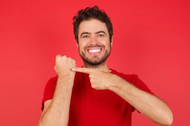 Kırmızı gömlekli yakışıklı beyaz adam, kol saatini göstermek için acele ediyor, sabırsız, rahat bir ifadeyle kameraya bakıyor.