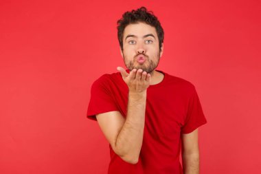 Tişört giyen yakışıklı beyaz bir adam izole edilmiş kırmızı arka planda kameraya bakıp havada öpüşürken güzel ve seksi görünüyor. Aşk ifadesi.