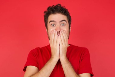 Neşeli, genç, yakışıklı, beyaz bir adam, izole edilmiş kırmızı arka plan üzerinde tişört giyiyor, neşe içinde kıkırdıyor, ağzını kapatıyor, tek başına eğleniyor, doğal kahkahalar atıyor, olumlu hikayeler dinliyor ya da komik anekdotlar dinliyor.