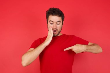 Şok olmuş genç, beyaz bir adam izole edilmiş kırmızı arka plan üzerinde tişört giyiyor. Şaşırtıcı bir şekilde aşağı bakıyor. Ön parmağıyla boş olduğunu gösteriyor. Korkmuş erkek, lüks mağazadan her zamanki gömleğin fiyatını öğrenir.