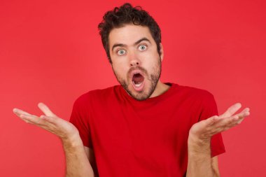 Hüsrana uğramış genç, yakışıklı, beyaz bir adam izole edilmiş kırmızı arka plan tişörtü giyiyor. Şaşırmış ve tereddütlü hissediyor, şaşkınlıkla omuz silkiyor, ağzını açık tutuyor, ne yapacağını bilmiyor..